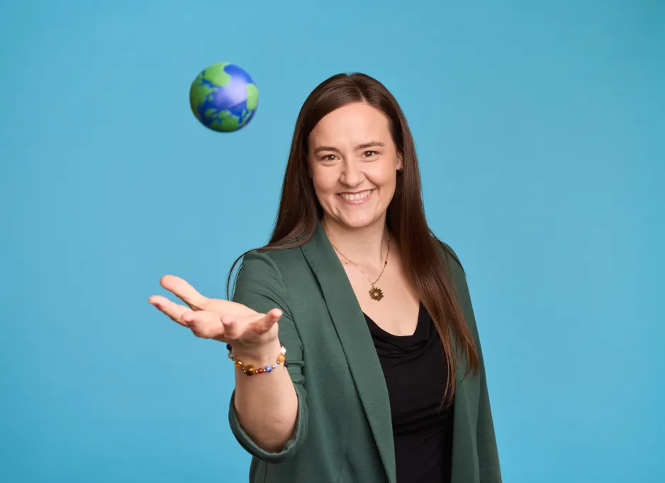 A smiling person with long brown hair and a green and black top stands in front of a blue background and throws a small ball decorated like the Earth up in the air