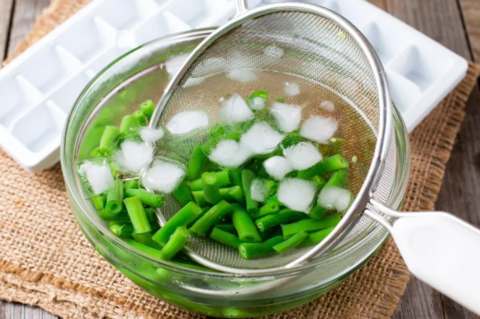 Des haricots verts coupés trempent dans un bol d’eau glacée et se font puiser avec une passoire.