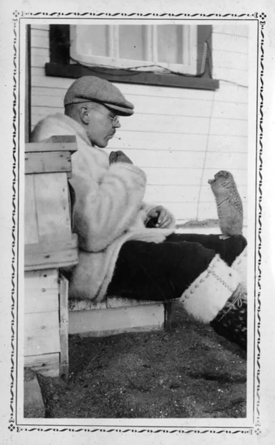 A black and white vertical photograph of a man seated in front of a white house, on the ground hand-feeding a squirrel that is standing on his leg reaching for food.