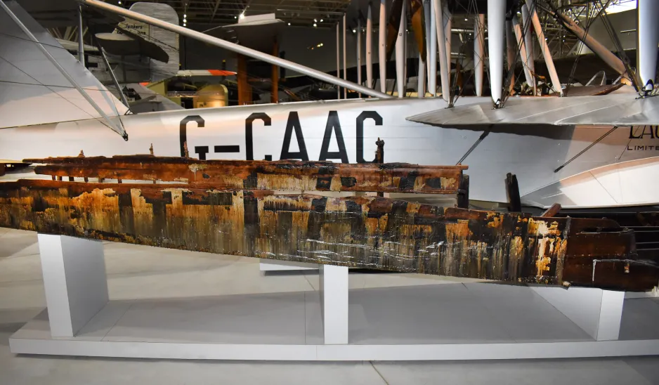 This image shows the starboard (right-side) tail sections of two HS-2L planes, one in front of the other. The one in the foreground is brown and wooden; on the surface there are big black letters, G-CAAC, which are faintly visible, and there is a section of missing material from the top half of the last C.