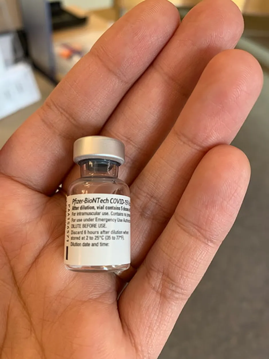 A tiny, glass vial sits in the palm of a hand. The words “Pfizer-BioNTech COVID-19” are visible.