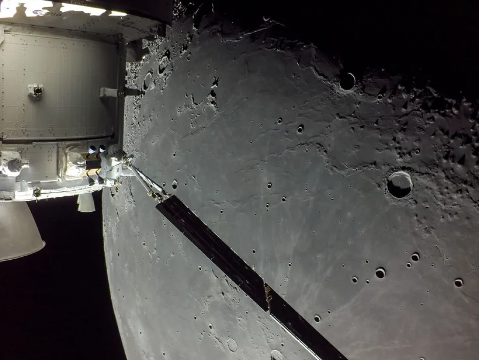A close up of the Orion spacecraft in the left foreground, and a portion of the lunar surface in the background. The surface is grey with a range of smooth and rough terrain and craters.