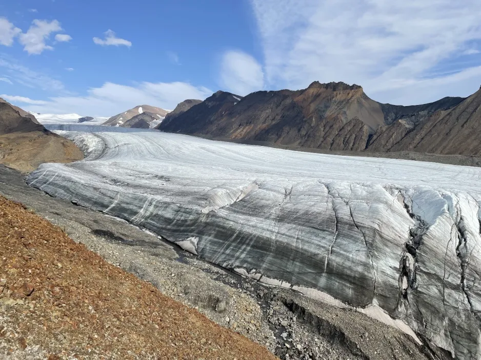 Vue en biais d’un grand glacier de vallée avec des bandes sinueuses qui alternent entre le blanc des gris foncés, et d’une plateforme de glace flottante supérieure au loin entourée de montagnes.