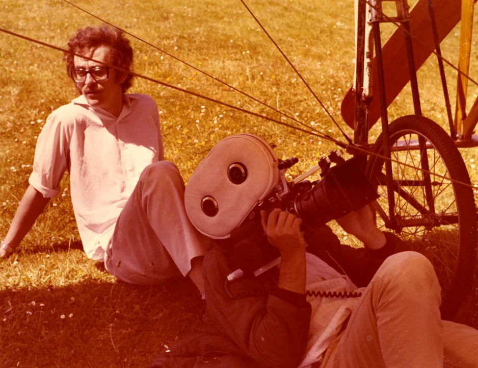 Les Harris sits back on the grass in the sun, while at work on the film Chabot Solo Part 1. He is sitting near a cameraman who is reclined on the ground underneath a Blériot XI aircraft, pointing an Arriflex camera up at the aircraft. The aircraft is mostly out of view, except for its wheel and part of its propeller.