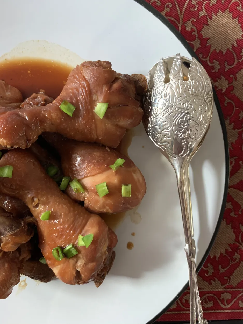 A close-up of a plate of medium-brown soy sauce chicken drumsticks. Green onion pieces are sprinkled on top, and a silver pair of tongs rests on the plate. A red floral placemat can be seen under the white plate.