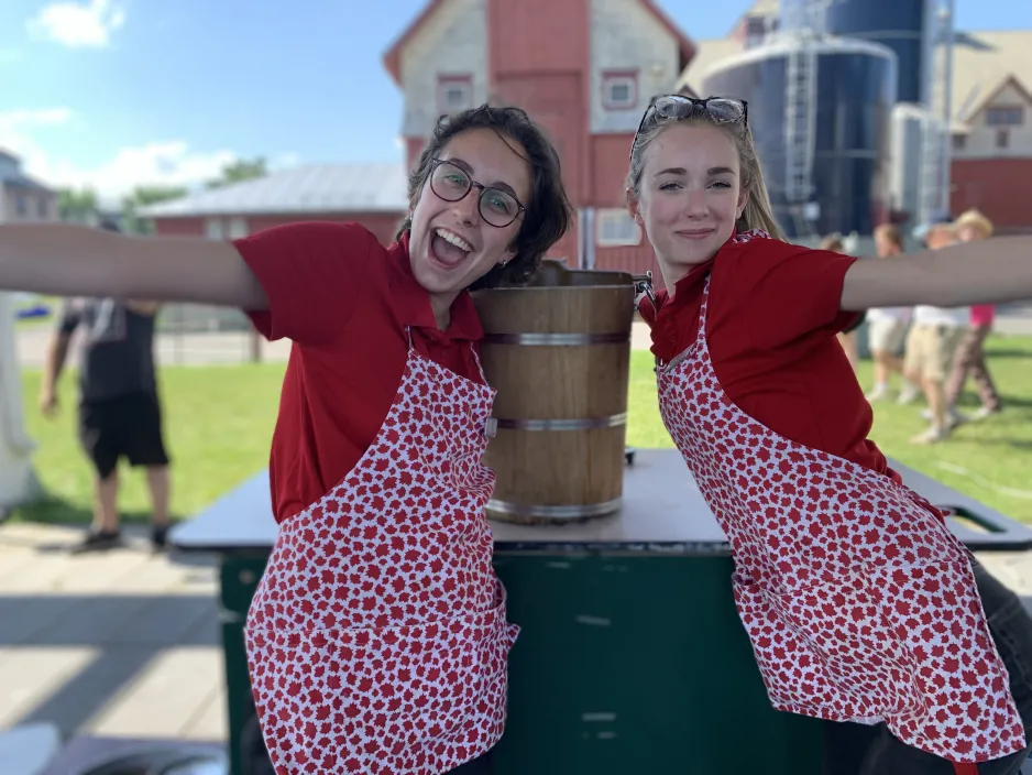 Two smiling people wearing red shirts and red and white aprons stand on either side of an old fashioned ice cream maker.  The museum's dairy barn is in the background.