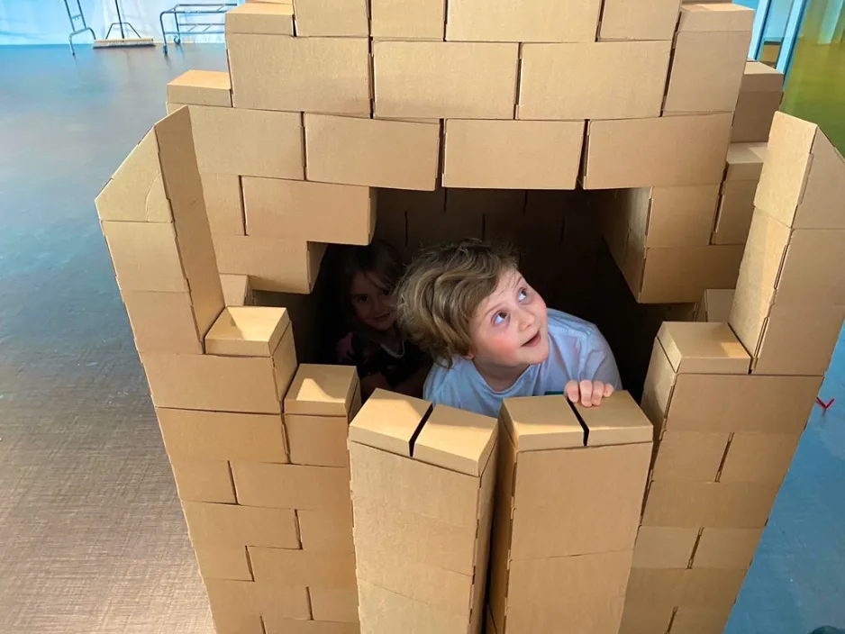 A child peaking outside of a small opening at the front of a house made out of cardboard blocks.