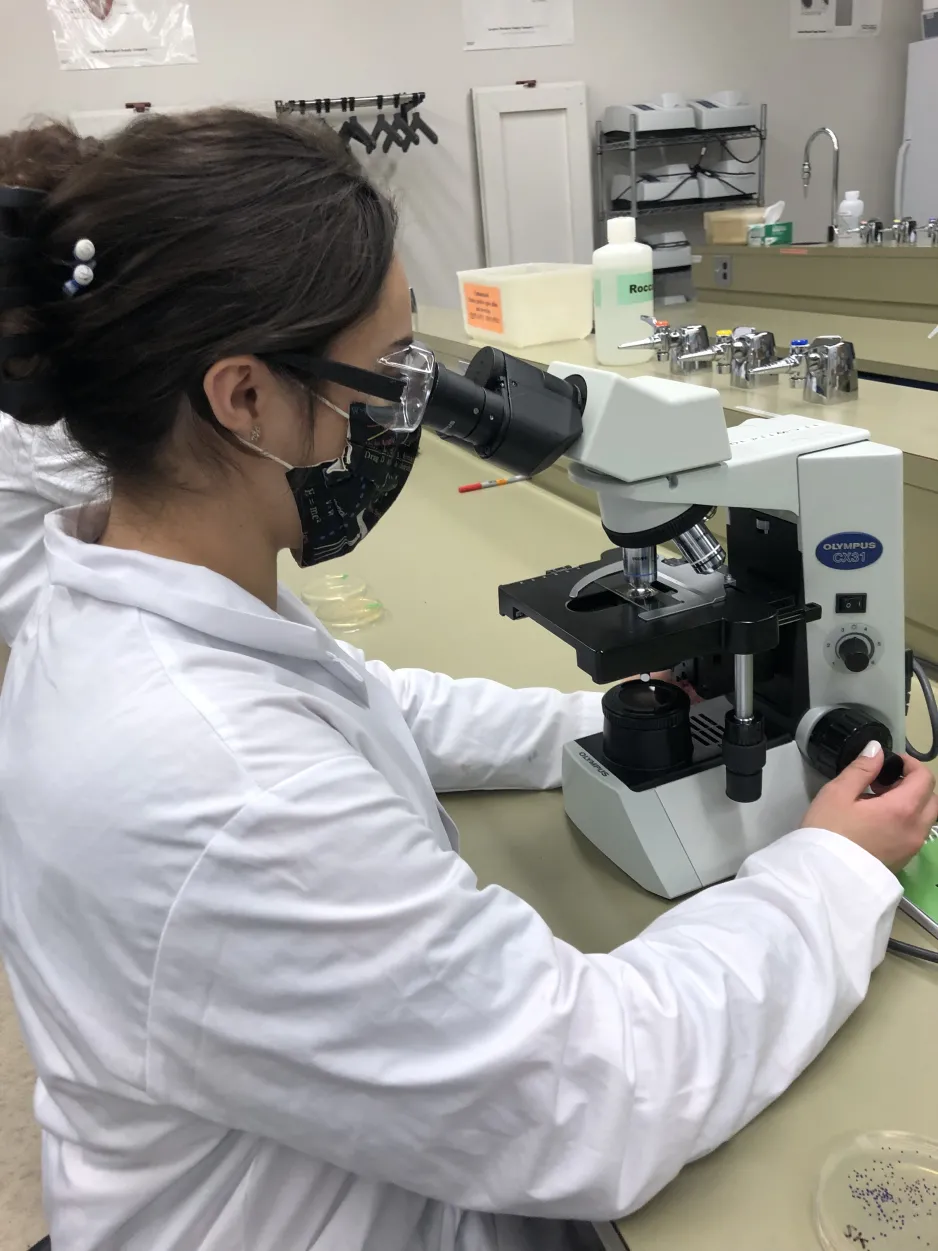 Samantha Burke looks through a microscope in a lab setting; she is wearing a white lab coat and safety glasses.