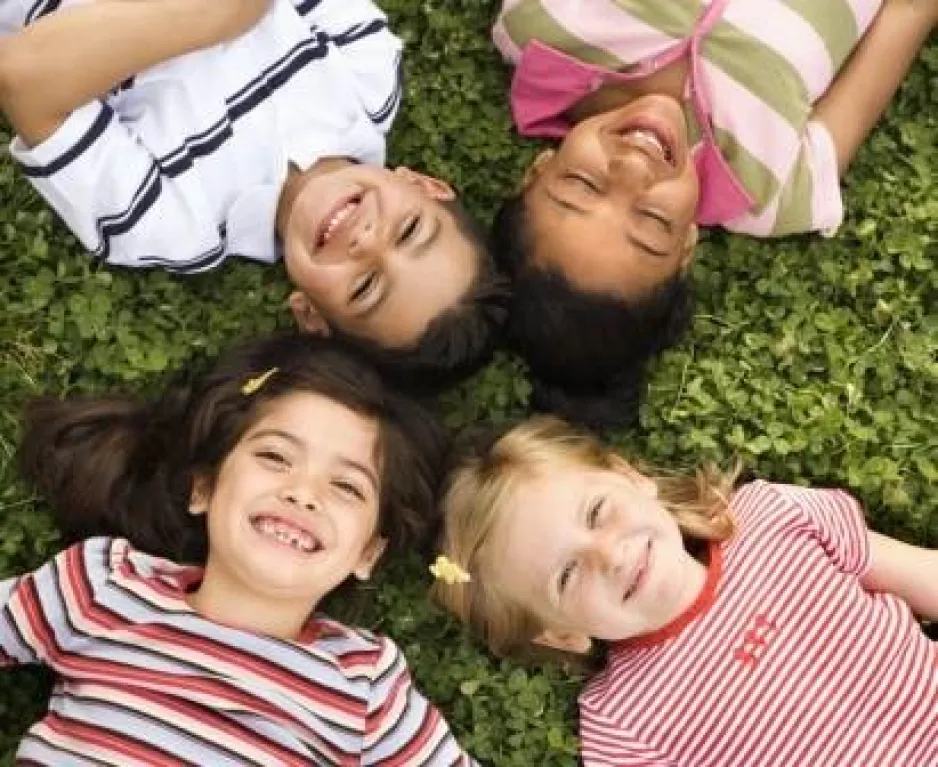 Vue de haut de quatre enfants souriants, allongés sur la pelouse, leurs têtes rapprochées au centre de la photo.