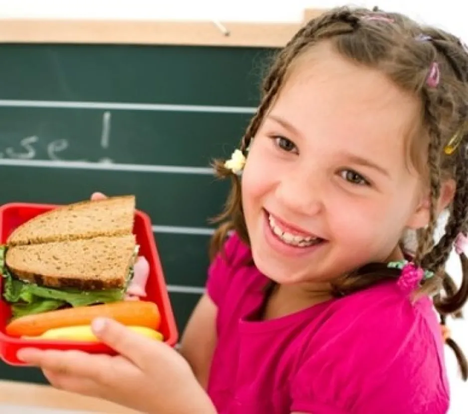 Une jeune fille souriante montre le contenu de sa boîte à dîner, qui contient un sandwich au pain à grains entiers et des carottes.