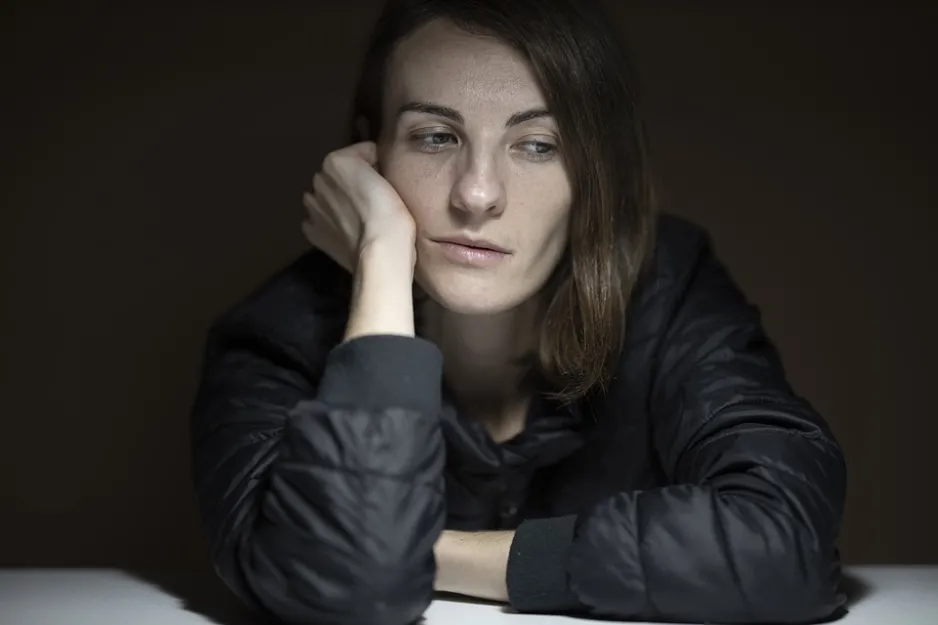 A seated women sits in a dark space, slouched over, resting her face on her hand. She is frowning slightly as she looks to the side, her eyes are partly shut, she has dark circles under her eyes, and she looks generally exhausted.