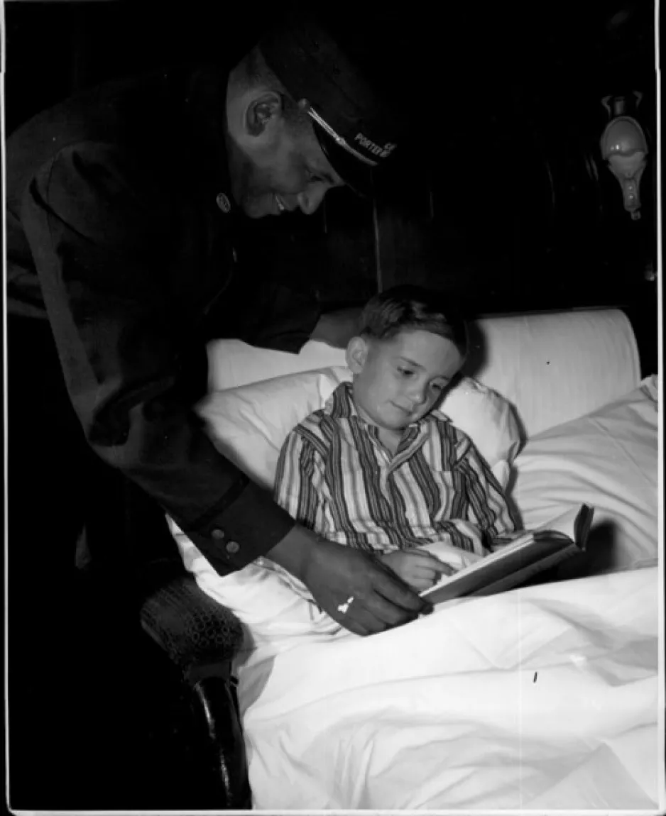 Black and white photograph, of a white child in bed with a book and a Black porter leaning over the child to help them.