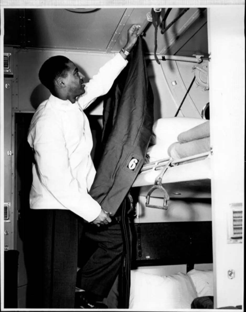 Black and white photograph of a Black porter standing on a ladder and pulling a curtain closed on an upper train berth.