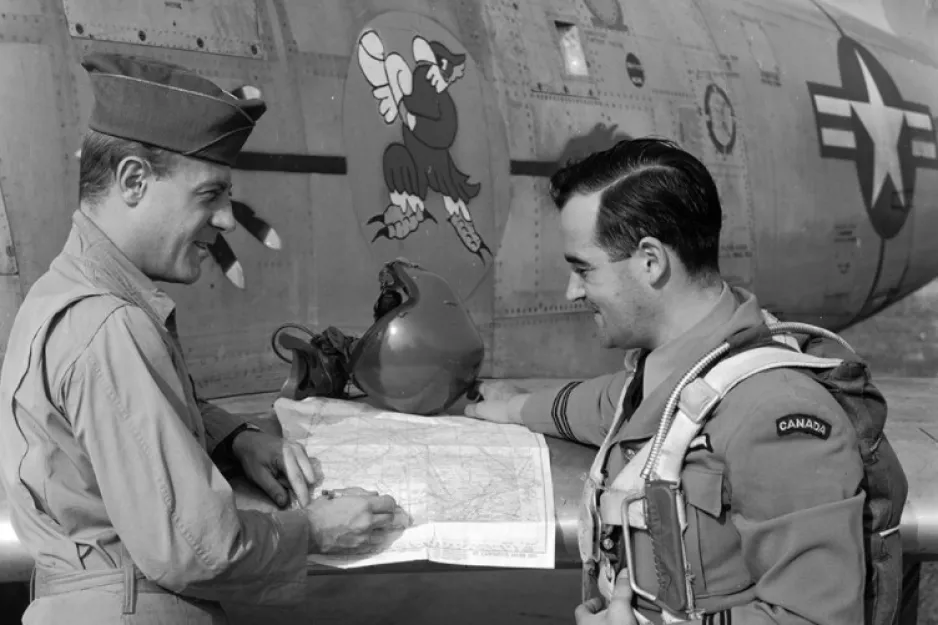 A black-and-white image depicts two men in military uniforms, standing next to an aircraft. Both men are in profile as they face each other. The man on the left is holding what appears to be a map.
