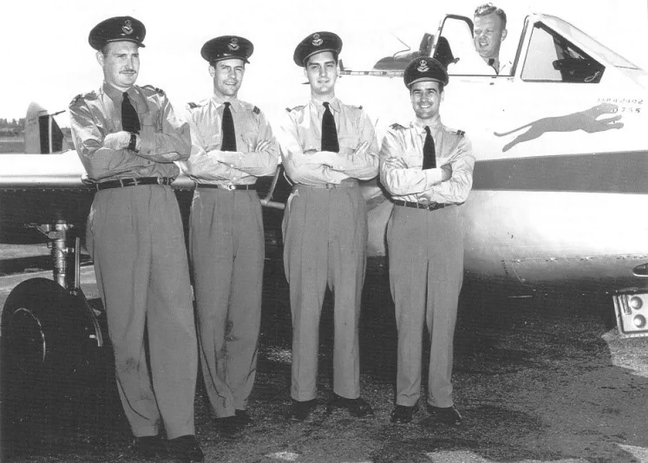 Une image en noir et blanc montre quatre hommes en uniforme militaire, posant devant un aéronef. Un homme se trouve dans la cabine de pilotage. Les quatre hommes sont debout un à côté de l’autre.