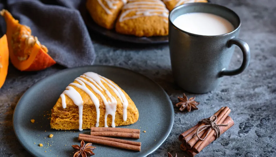 A scone sits on a plate, along with some cinnamon sticks and spices. In the background, another plate of scones, some pumpkin slices, and a cup of milk are visible.