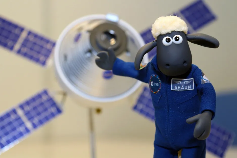 Shaun, le mouton-jouet, vêtu d’une combinaison de vol bleue est en avant-plan et un petit modèle de l’astronef Orion se trouve en arrière-plan.