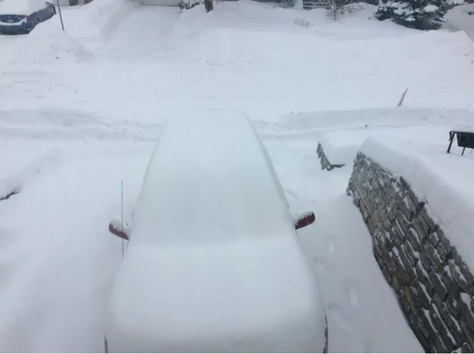 Image is a photograph of a parked car in a snowy driveway, with about 30 cm of snow on top of its hood.  