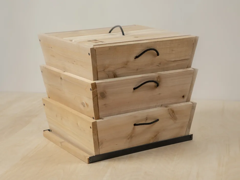 Three light-coloured, cedar boxes are stacked on top of one another on top of a light-coloured surface. Each box has a contrasting black loop handle in the front, and another black loop is visible on the lid of the top box.