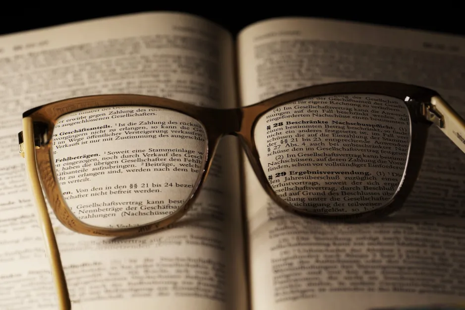 Une paire de lunettes brunes reposent sur le dessus d’un livre ouvert. Le texte que l’on voit à travers les lunettes est clair, le reste est flou. 