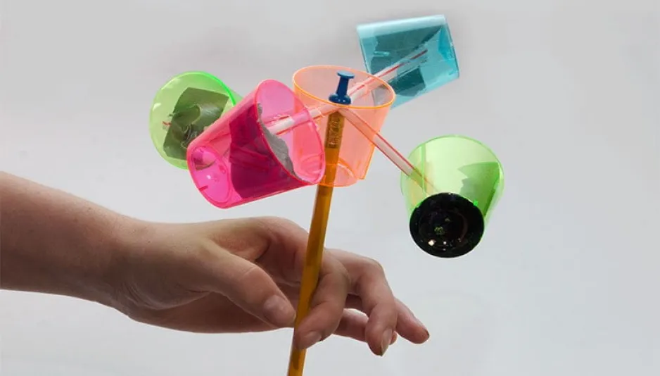 Une main tient un engin composé d'un crayon, d'une punaise, de pailles et de cinq gobelets de plastique colorés. 