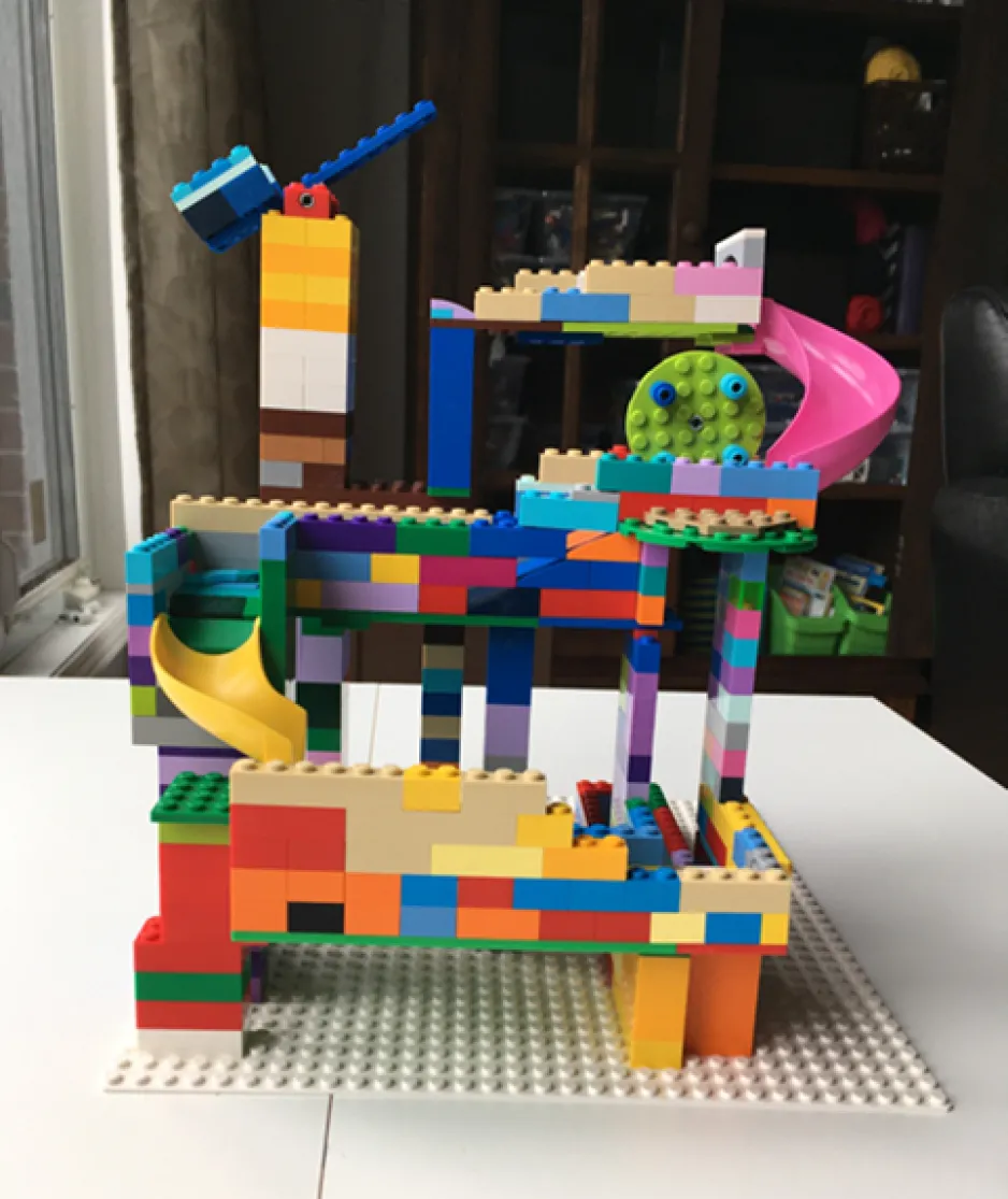 Une image d’une création LÉGO avec un levier et des glissades sur des différents niveaux. Elle est faite de blocs multicolores. La création est debout sur une table blanche et une bibliothèque dans l’arrière-plan.
