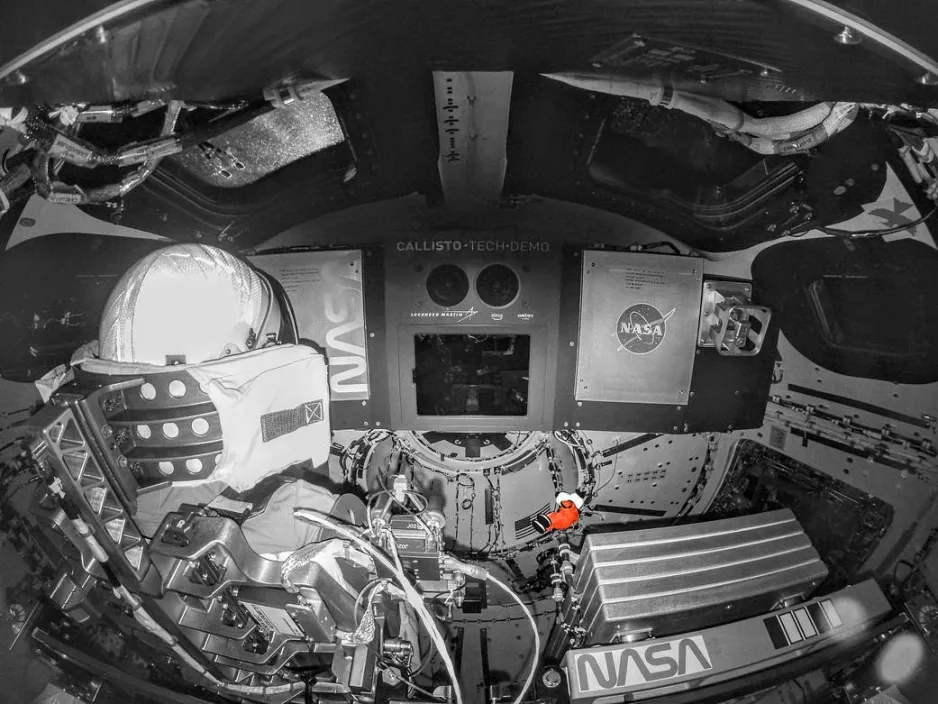 Greyscale view inside the Orion spacecraft busy with tubes, knobs, and cables. There are windows above, a manikin seated on the left, a Callisto tech demo in the centre with a small coloured Snoopy doll with orange spacesuit floating in the centre.