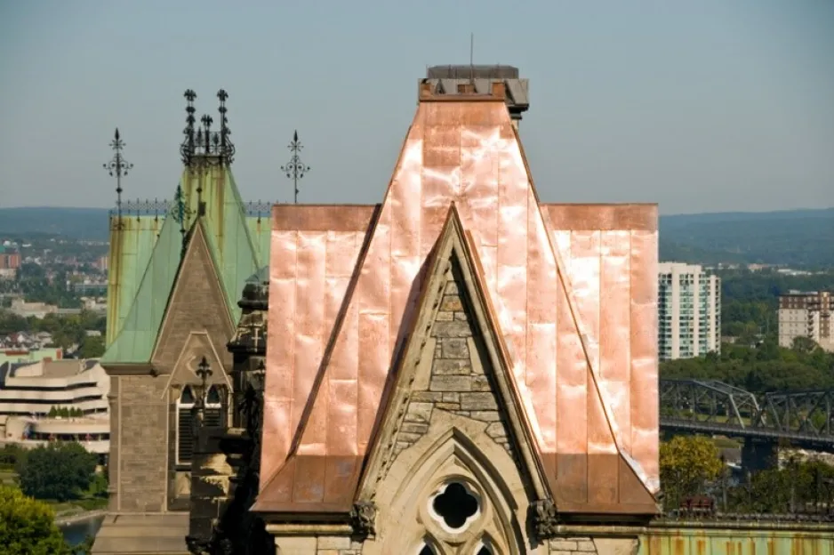Gros plan d’une section du toit de l’édifice Ouest du Parlement canadien. En avant-plan, on remarque une partie en cuivre neuf et brillant, alors qu’au milieu, on observe une partie en cuivre vert vieilli. En arrière-plan, on aperçoit une vue aérienne de la rive de Gatineau.