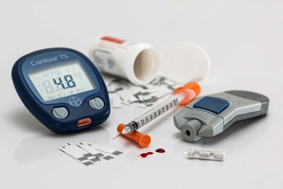 Blood glucose test strips and a digital blood glucose monitor lie on a white surface. An insulin needle lies in the centre, with a couple drops of blood lying under the needle tip. 