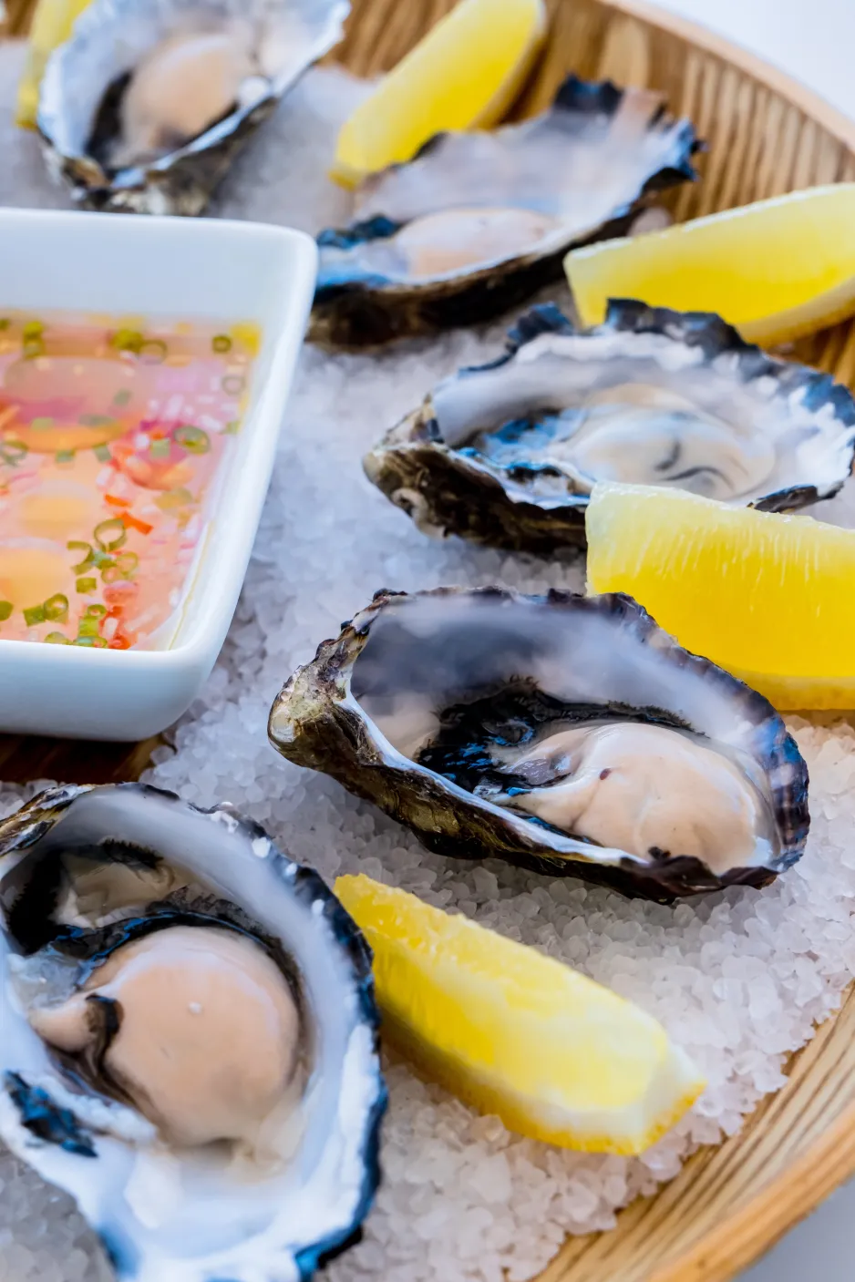 In a shallow wooden bowl, shucked oysters and lemon wedges are laid out in a circular fashion on a bed of coarse salt.