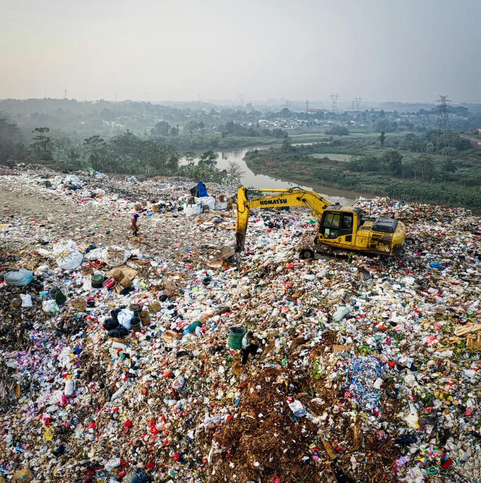 an excavator sits on top of a landfill, surrounded by trash. In the background is a river flowing into a city.  