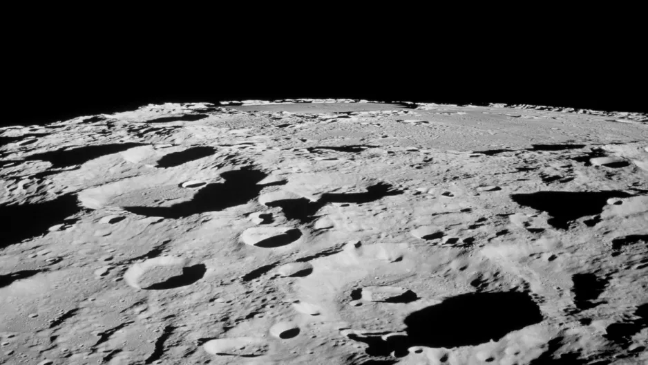 Close-up view looking at the lunar surface at an angle. The Moon’s surface is bright gray and riddled with round depressions in a range of sizes, set against the black background of space.