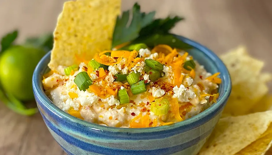 A bowl of dip sits on a wooden tabletop. Tortilla chips, green onions, and cheese are visible on top of the dip, and some chips and a lime are next to the bowl. 