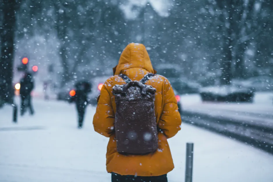 A rear view of a person wearing a brown backpack and a thick yellow coat with the hood up. They are walking down a snow-covered street with cars and other pedestrians in the background. It is snowing heavily. 