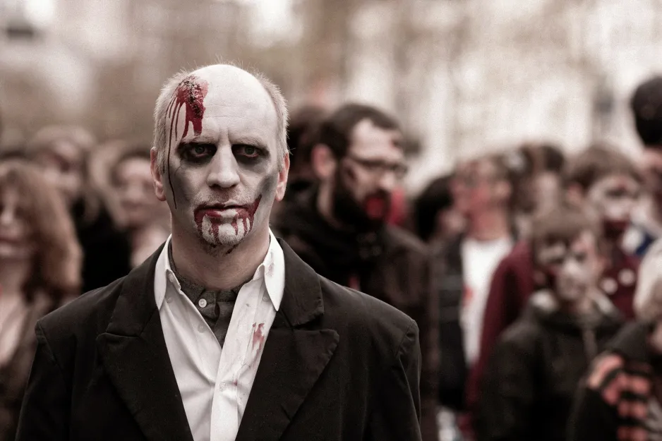 A man dressed up as a zombie wears a black suit, white face paint, and fake blood on his head and around his mouth. In the background are a group of people also dressed up as zombies. 
