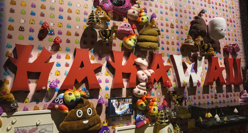 A bunch of plush poomojis and a few golen poomojis attached to a wall with pink paper with an array of colourful poomojis on it. On the wall it says "Kakawaii." 