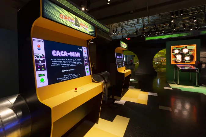  Deux jeux d'arcade avec des manettes de jeu côte à côte et un pinball à droite. Le jeu à l'avant-plan porte l'inscription « Caca-Man.»