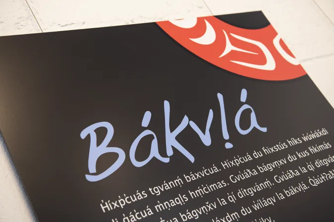 Un gros plan d’une affiche sur laquelle on peut lire le mot « Bákvḷa » en grosses lettres bleues sur un fond noir. On peut voir un élément décoratif rouge dans le coin supérieur droit de l’affiche avec du texte blanc en langue Heiltsuk sous le titre principal.