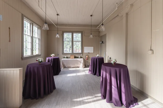 Une grande pièce avec des murs blancs et un haut plafond blanc dans une grange rustique très propre. Plusieurs tables hautes sont disposées le long des murs, drapées de nappes violet foncé.