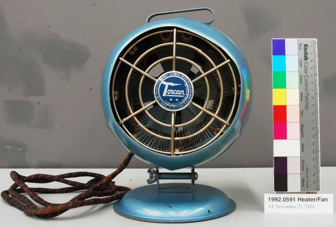 The front view of blue electric heater, with a dark blue label that says “Torcan” in the middle. A black-and-red braided cord sits to the left of the fan. 