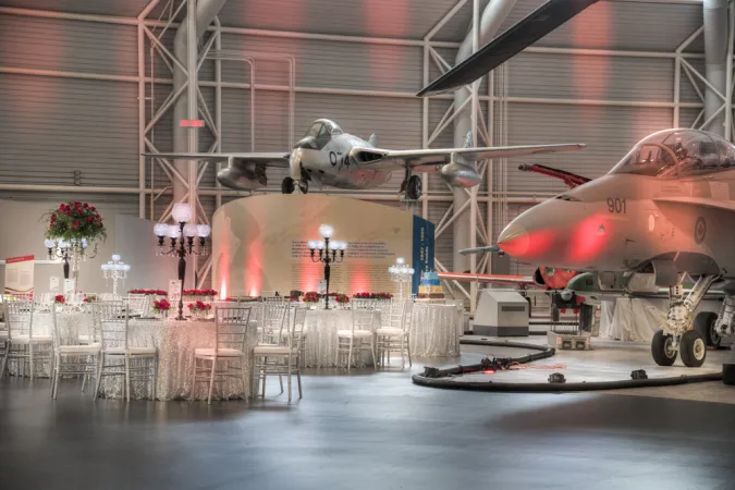 A view inside the Canada Aviation and Space Museum with two white airplanes in the background. Large tables are set up around the room with white translucent shimmering table cloths and elegant white chairs. Scattered around the room are classical Victorian-style lampposts with round lamp bulbs with geometric patterns on the glass