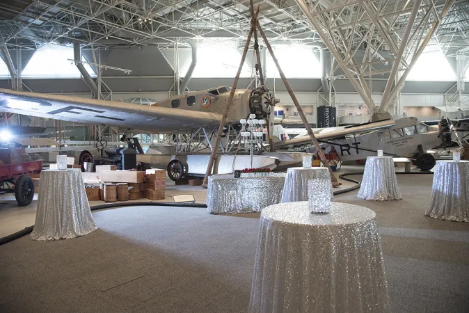Inside the Canada Aviation and Space Museum. Several shimmering white tablecloths are draped over small high round tables with white shimmering vase centrepieces at each table. There is a white lamppost near the tables with an airplane in the background. White support beams are in the background blending into the white walls and ceilings. 