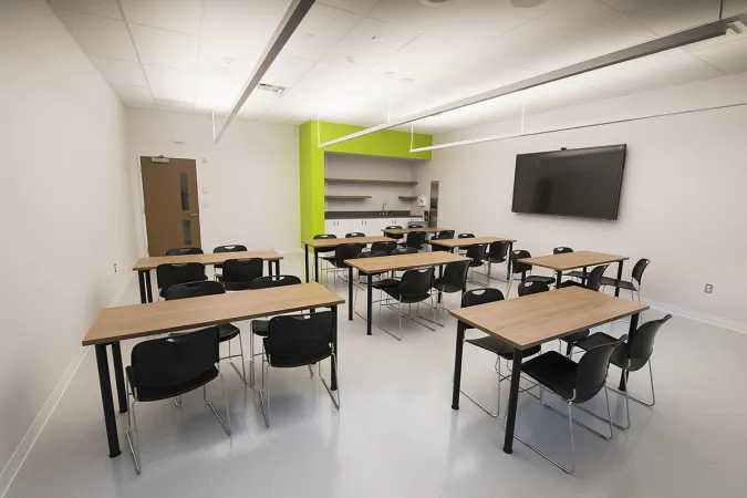 Une salle de cours spacieuse avec une télévision sur un côté et huit tables avec quatre chaises chacune qui occupent l’espace. Sur un autre mur se trouvent un évier et quelques étagères.