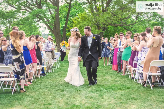 Beautiful outdoor wedding ceremony on a lush green lawn under large trees. Rows of people are standing and clapping. A bride and groom are walking down the centre aisle holding hands and smiling.