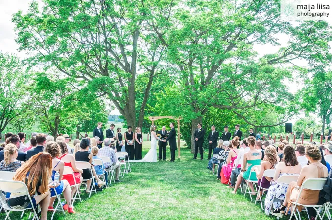 Beautiful outdoor wedding ceremony on a lush green lawn under large trees. Rows of people are sitting on white foldout chairs and a bride and groom are standing at the front under an arch with an officiant.
