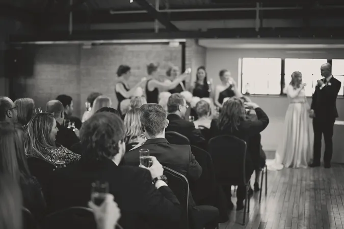 A black and white rendering of a photo of a bride and groom standing at the front of the room at a microphone. Many people are seated in rows watching the two. Bridesmaids are lined up at the front of the room.