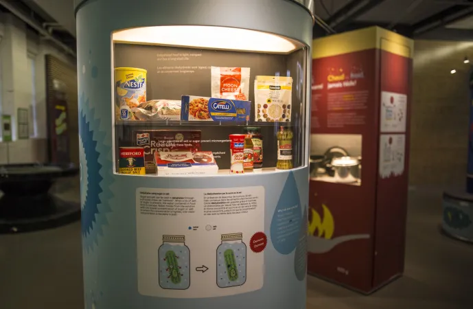 And circular exhibition display case with different food packaged, and graphics of jars with liquid in them can be seen in the foreground. Another display can be seen in the background.