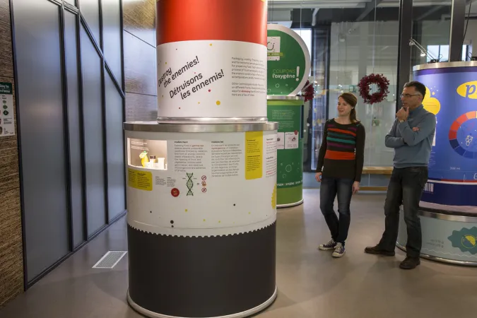 Two people are reading texts on a cylindrical exhibition model with the title “Destroy the enemies!”  Other exhibition modules are behind them.