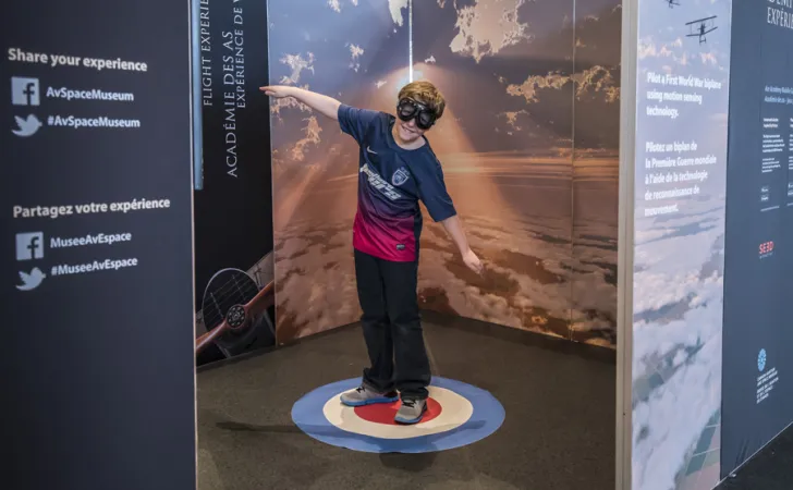 A child wearing a blue and red t-shirt, black pants and aviation goggles. Stands on a bulls-eye carpet with their arms out.  Panels behind them are of the sky above the clouds. Foreground panel has social media information.