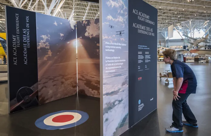 A child leans in to read text on a large exhibition panel.  Behind this panel are two more panels.  The one to the left has the exhibition title Ace Academy Flight Experience running from bottom to top.  The panel on the right has an image of the sky above the clouds.